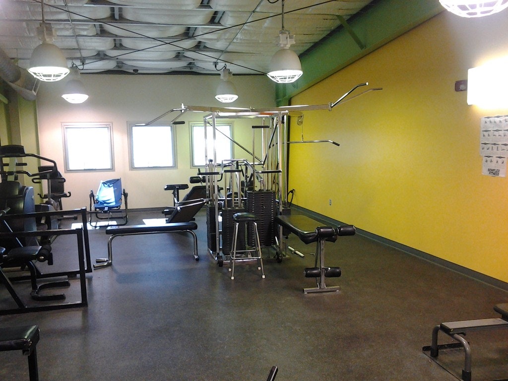 An empty gym with various workout machines, green and yellow walls, and overhead industrial lighting.