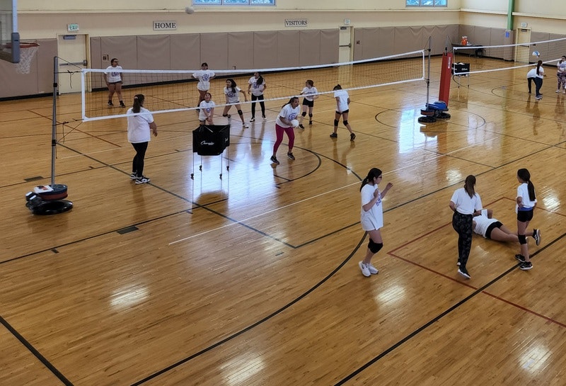 An indoor volleyball court with players practicing and a robotic vacuum cleaner on the floor.