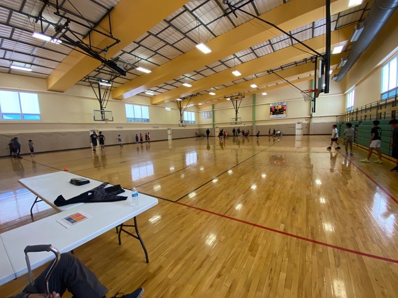Indoor basketball court with players, scoreboards, a table with belongings, and spectators on bleachers.
