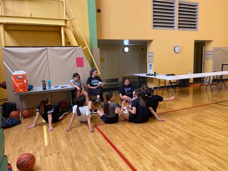 A group of girls in sportswear resting in a gym with a basketball, water cooler, and folding table visible.