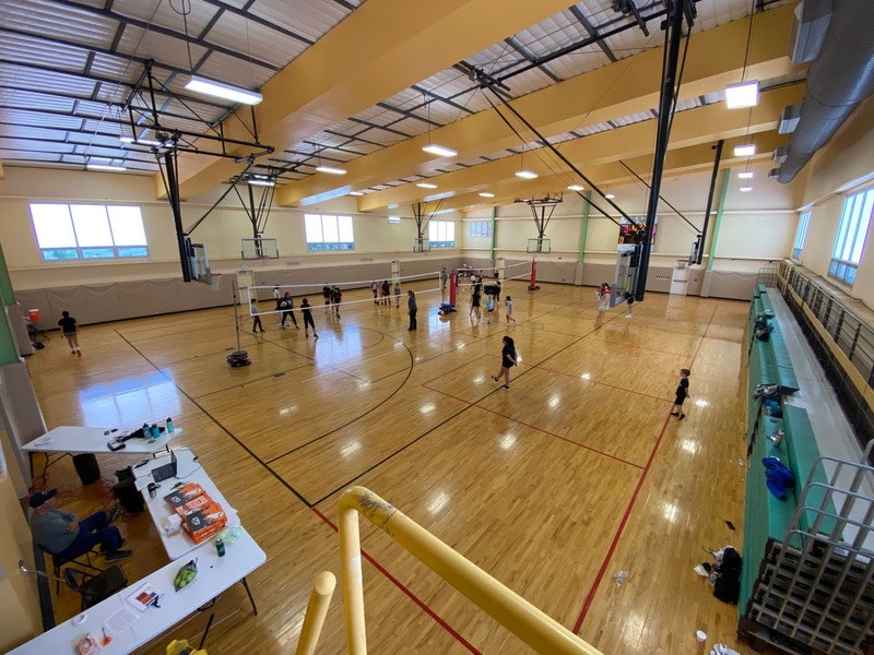 Indoor gym with people playing volleyball, basketball hoops, bright lighting, and bleachers.