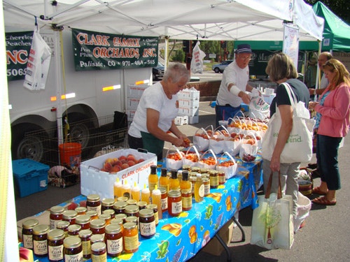 A farmers' market stall with fresh produce and jams, and people browsing or buying items.