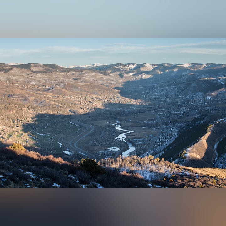 A valley with a meandering river, roads, buildings, surrounded by snow-dappled mountains under a clear sky.