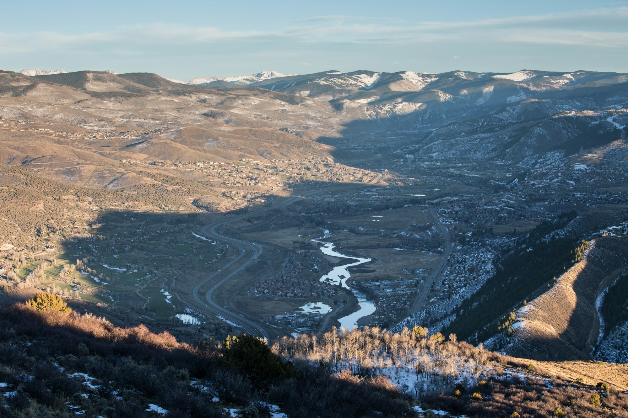 A valley with a meandering river, roads, buildings, surrounded by snow-dappled mountains under a clear sky.