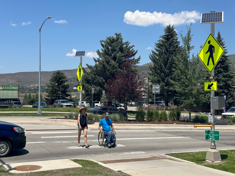 Two people at a crosswalk, one in a wheelchair, solar panels on signs, sunny day.