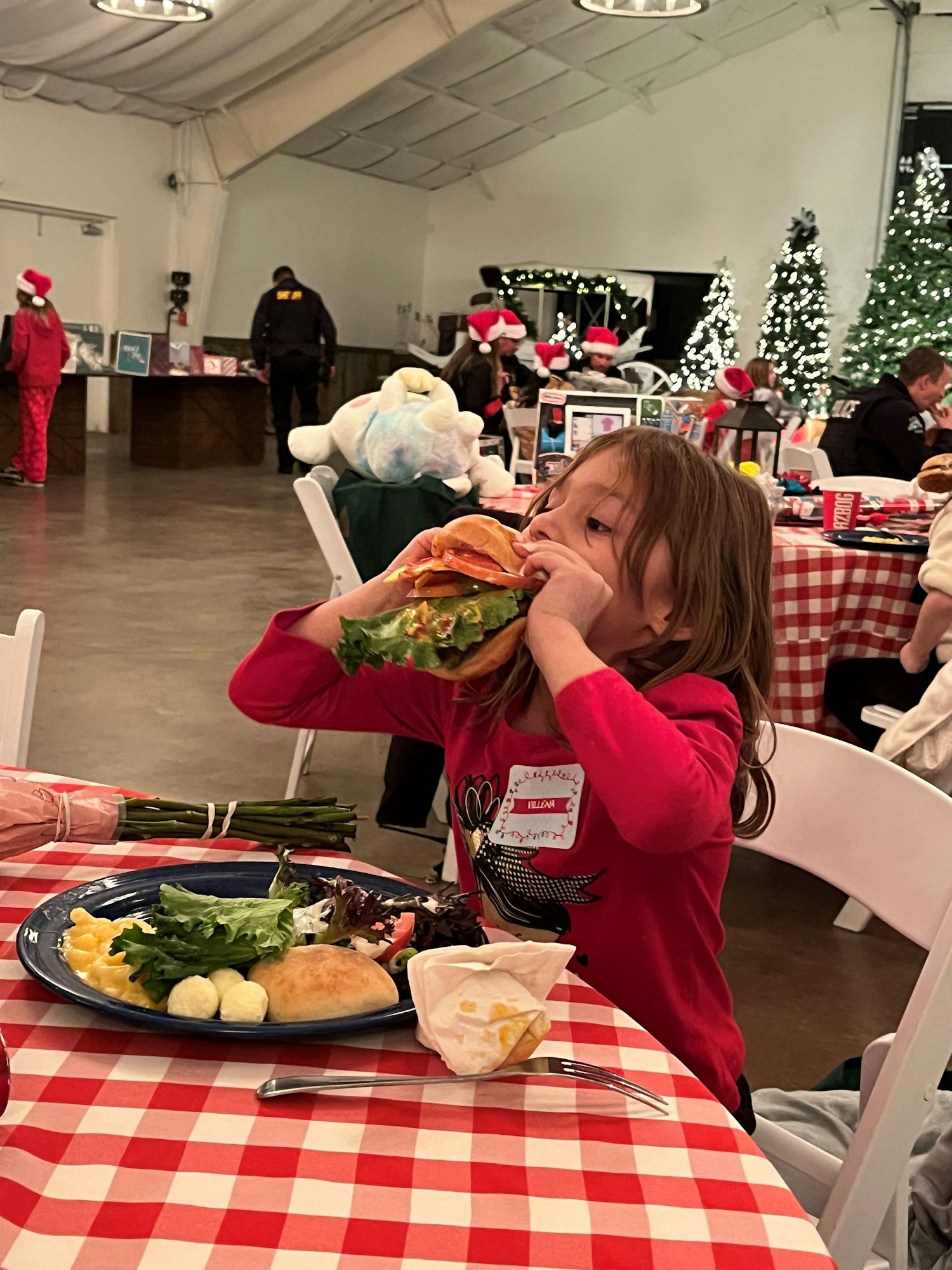 A child in red eats a big sandwich at a festive event with decorated trees and people wearing Santa hats.