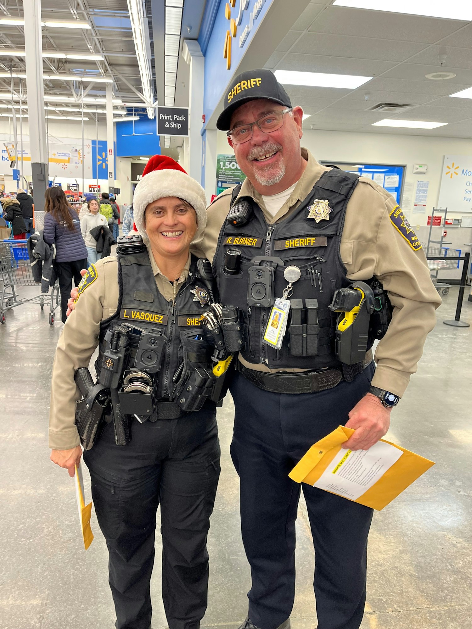 Two smiling sheriffs in uniform at a store, one wearing a Santa hat.