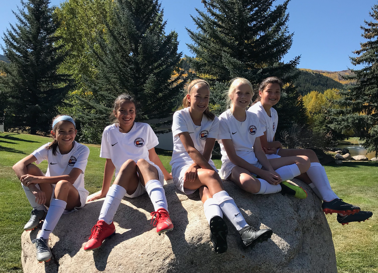 A group of young soccer players in uniform sitting on a rock with trees and autumn foliage in the background.
