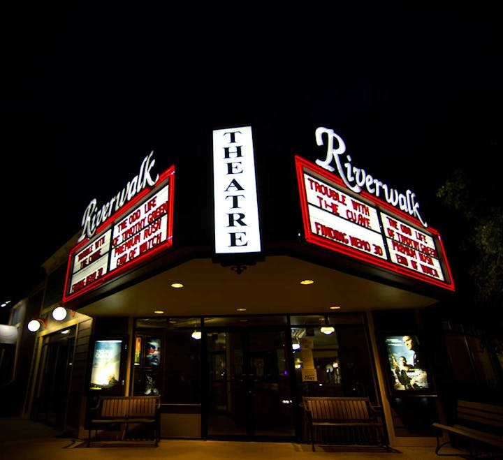 A night view of the Riverwalk Theatre marquee illuminated with movie titles.