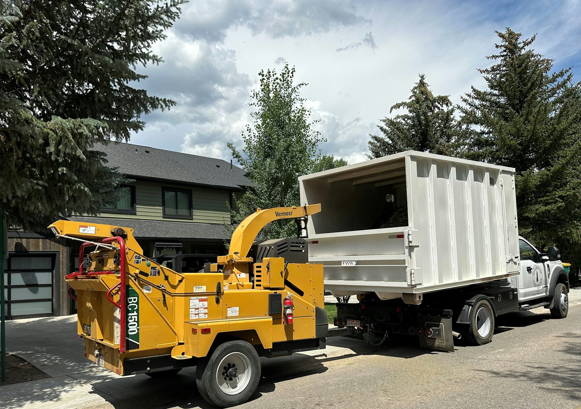 A wood chipper attached to a truck with an empty container, likely for collecting wood chips, parked on a residential street.