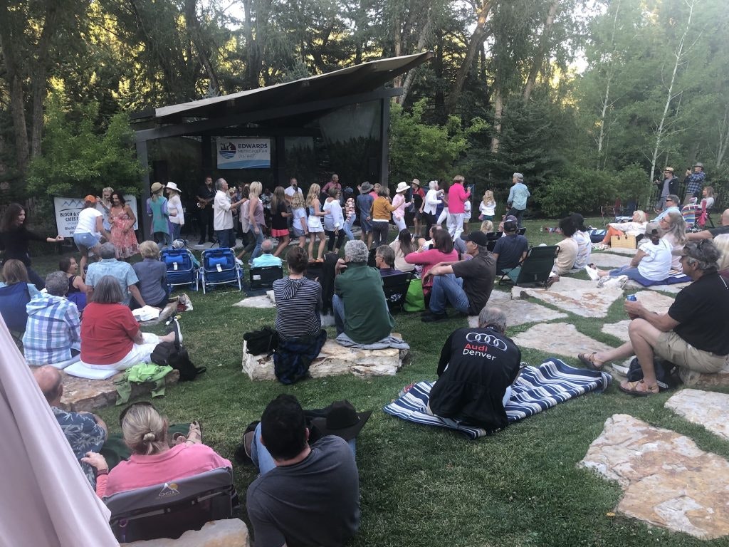 People gathered outdoors, sitting on lawns and rocks, watching a performance on stage in a park-like setting.