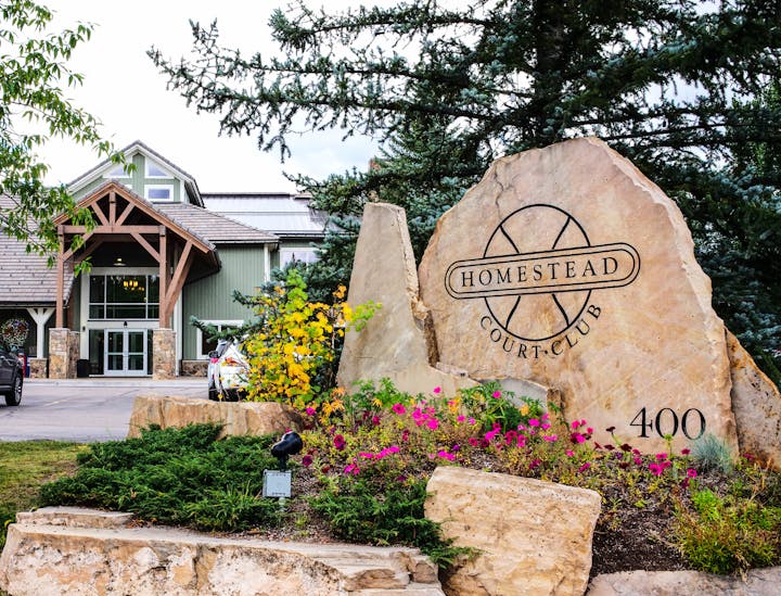A building with a sign "HOMESTEAD COURT CLUB" on a large stone, landscaping with flowers, and a clear sky.