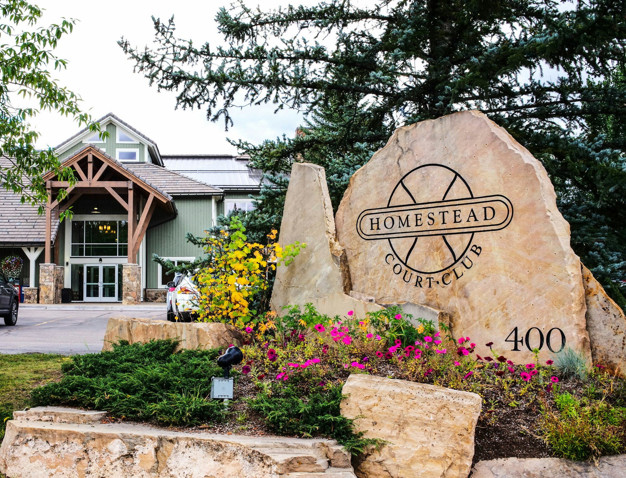 A building with a sign "HOMESTEAD COURT CLUB" on a large stone, landscaping with flowers, and a clear sky.