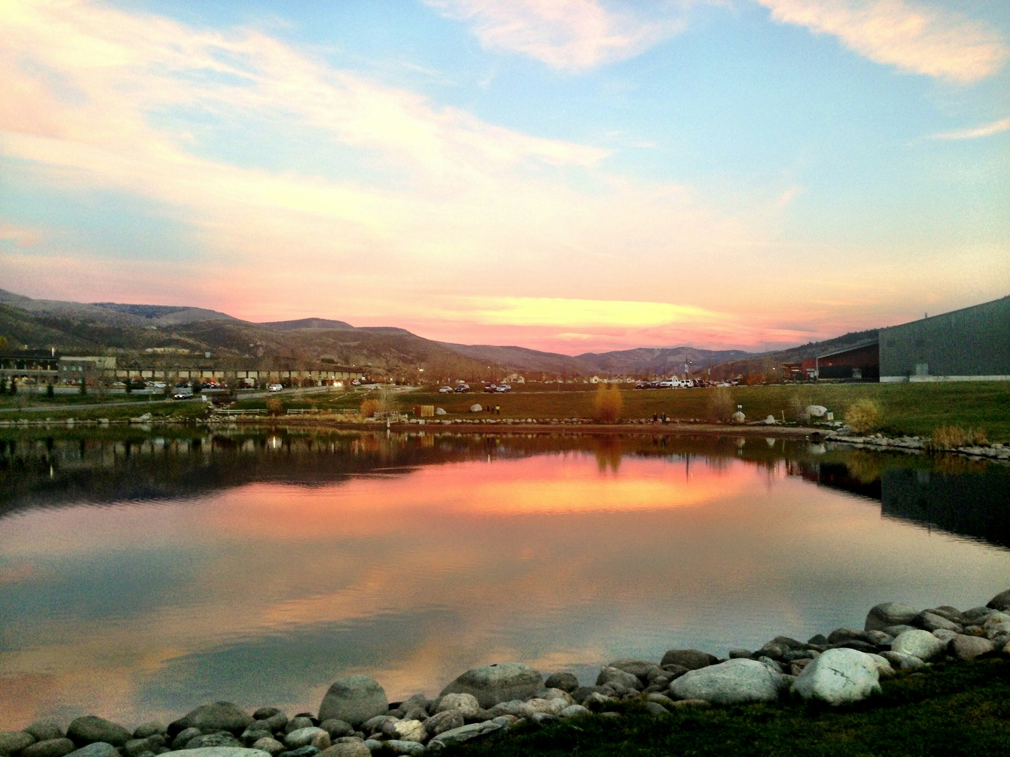 Sunset reflected in a pond with rocky shore, industrial buildings aside, under a pastel sky.