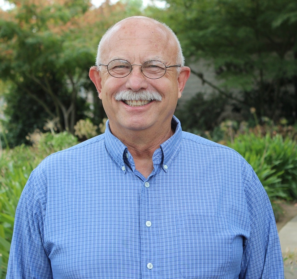 A smiling man with glasses and a mustache, wearing a blue checked shirt, with greenery in the background.