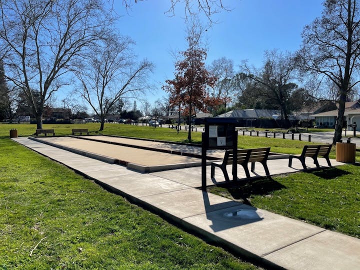 A bocce ball court in a park with benches, trees, and a clear sky.