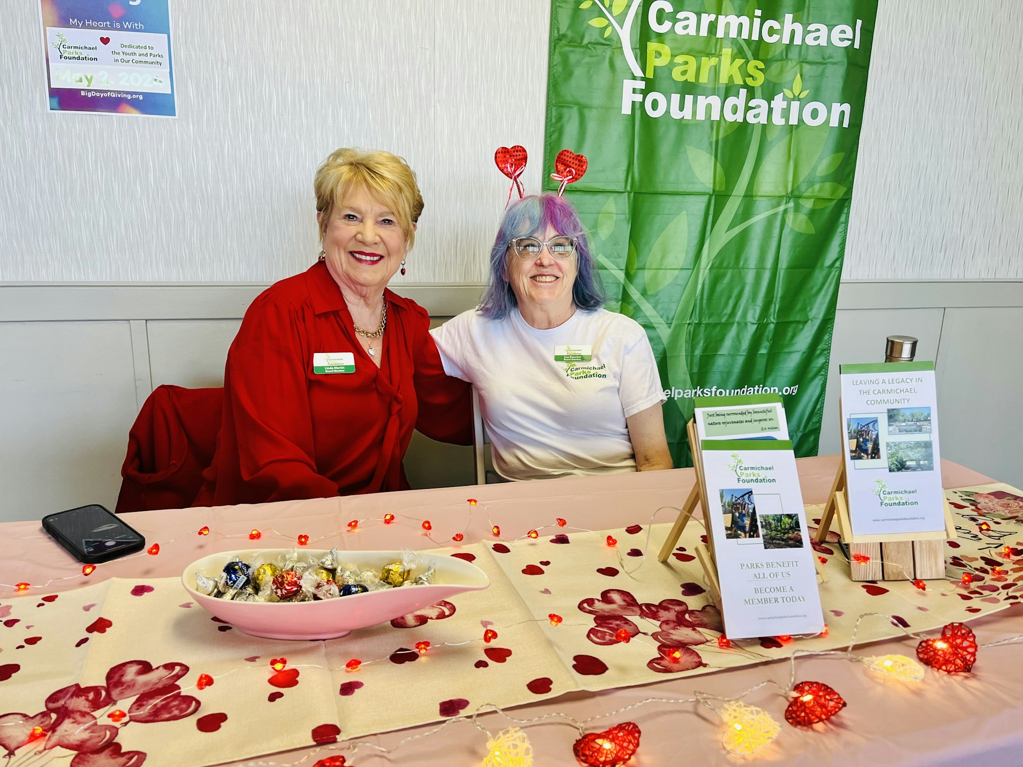 Two women at a Carmichael Parks Foundation booth with promotional materials and candy on a decorated table.