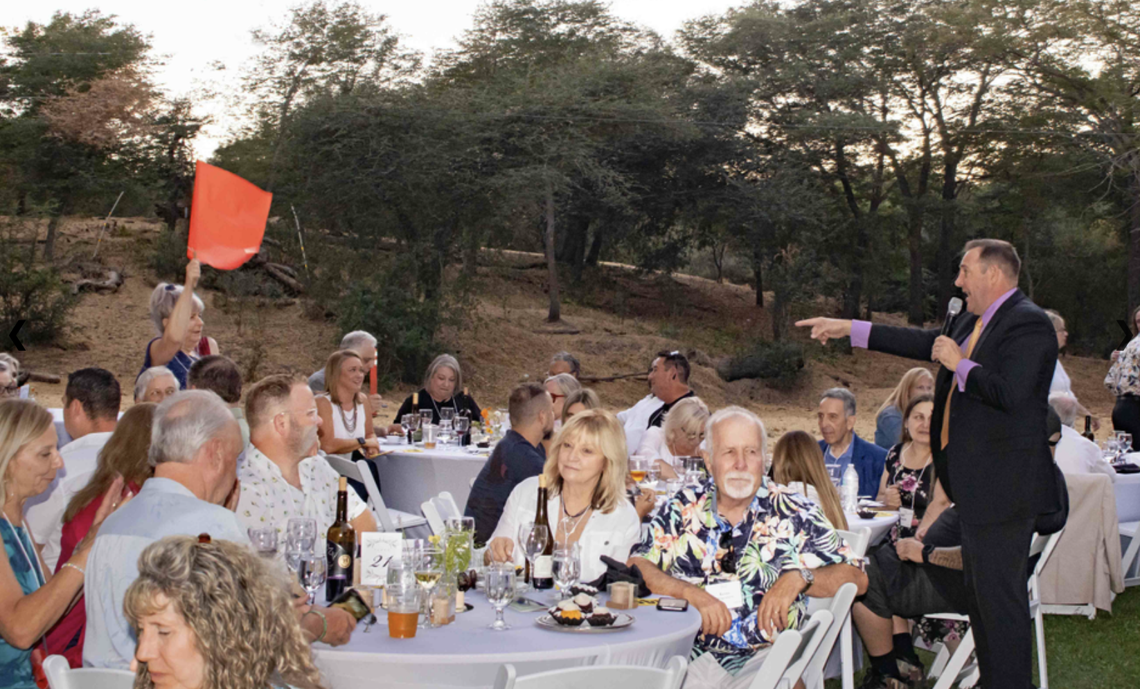 Outdoor event with people seated at tables, a woman holding a red flag, and a man speaking with a microphone.