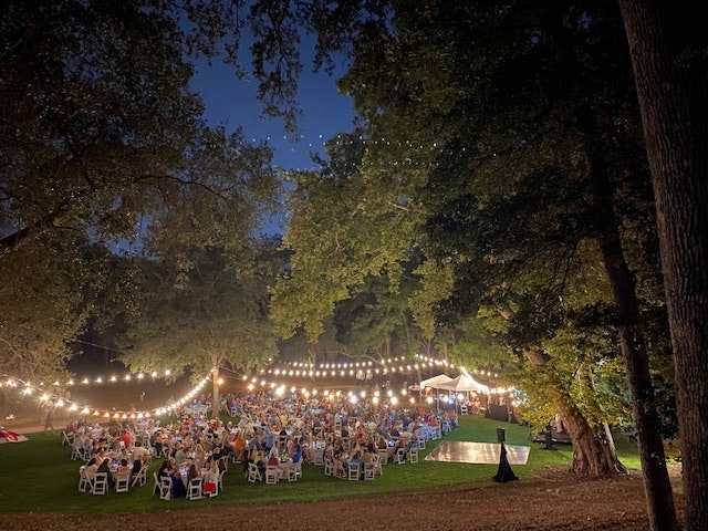 Outdoor evening event with people seated at tables, string lights, trees, and a stage.