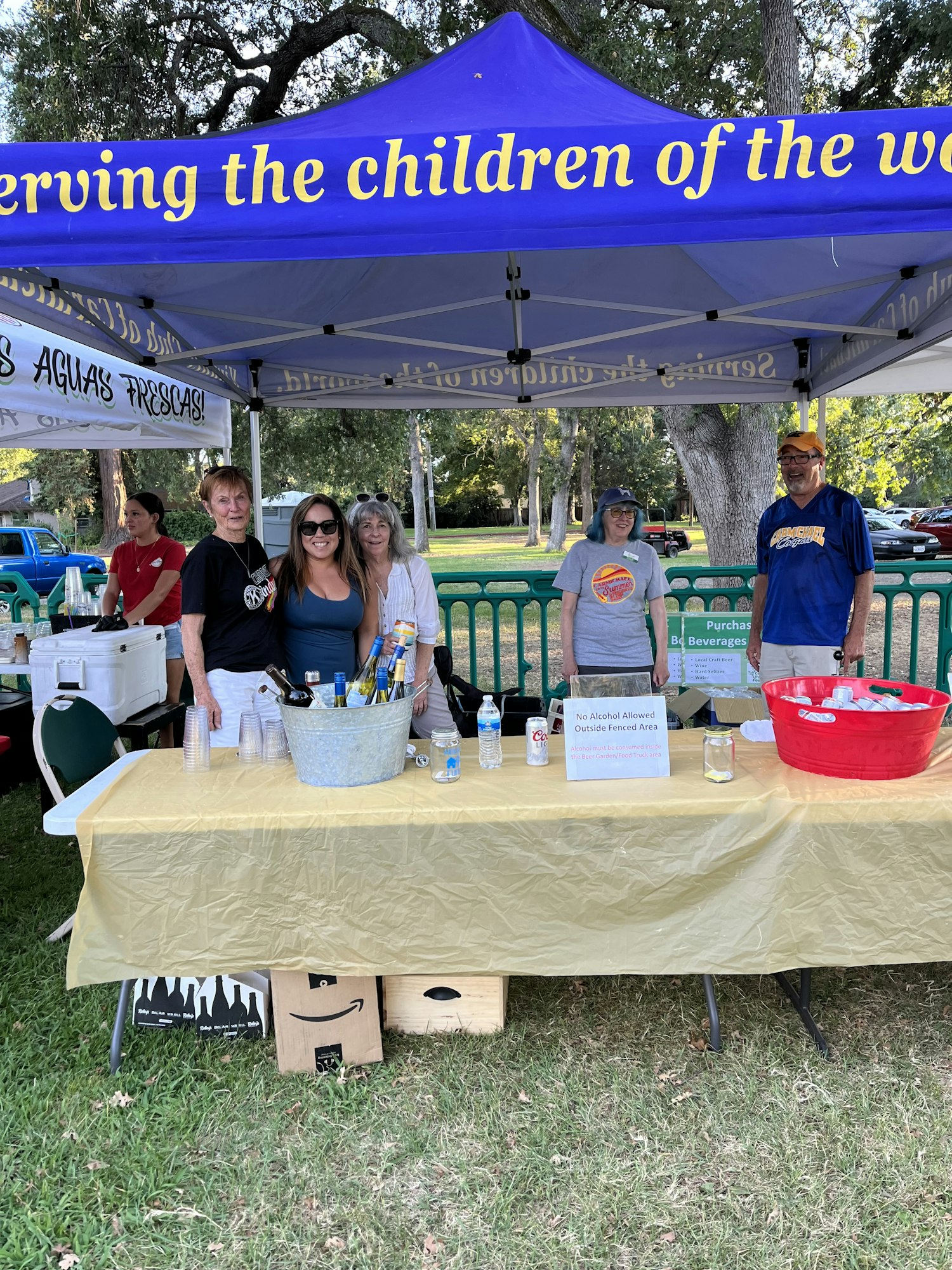 Outdoor event with people by tables under tents, text saying "Serving the children of the world." Bottles of water on table.