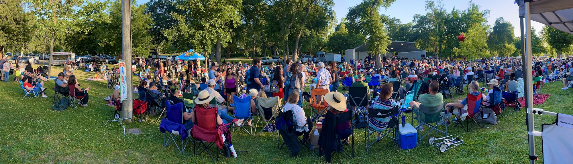A large outdoor gathering with people seated on chairs and picnic blankets in a park, surrounded by trees and a festive atmosphere.