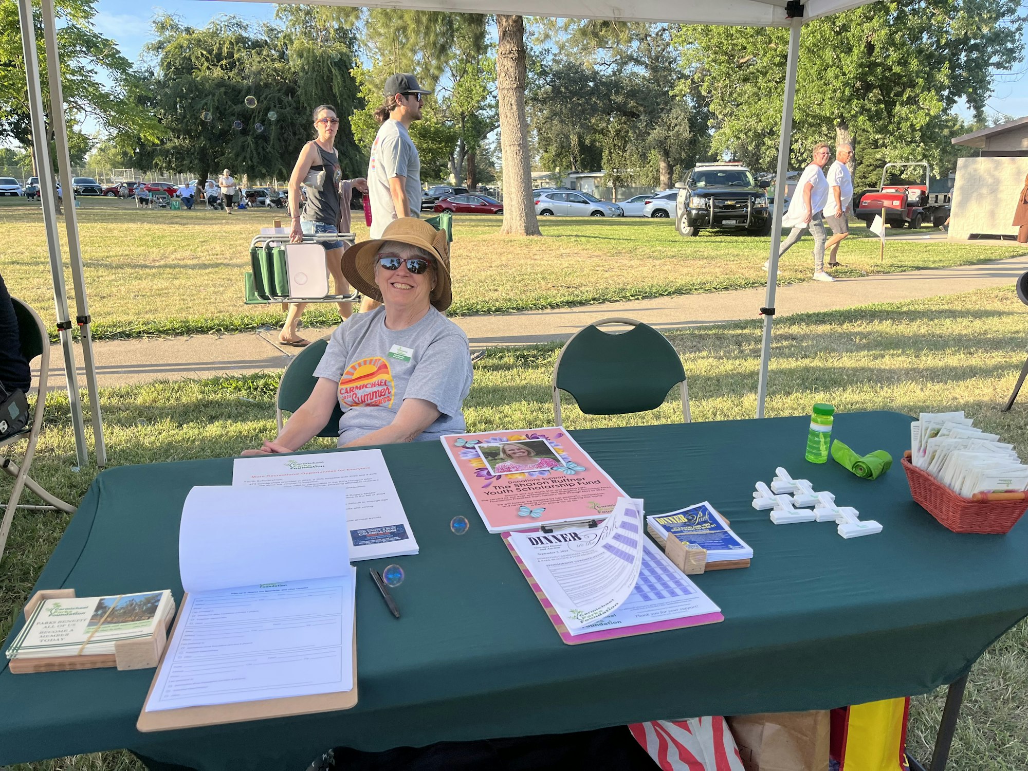 A smiling person at an outdoor info booth with papers, flyers, and a sign-up sheet on a sunny day.