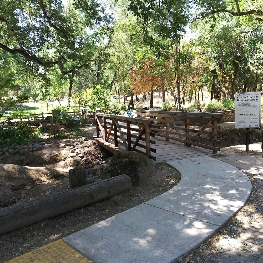 A wooden bridge in a park with trees, a sign for Charles C. Jensen Botanical Garden, and a person in the background.