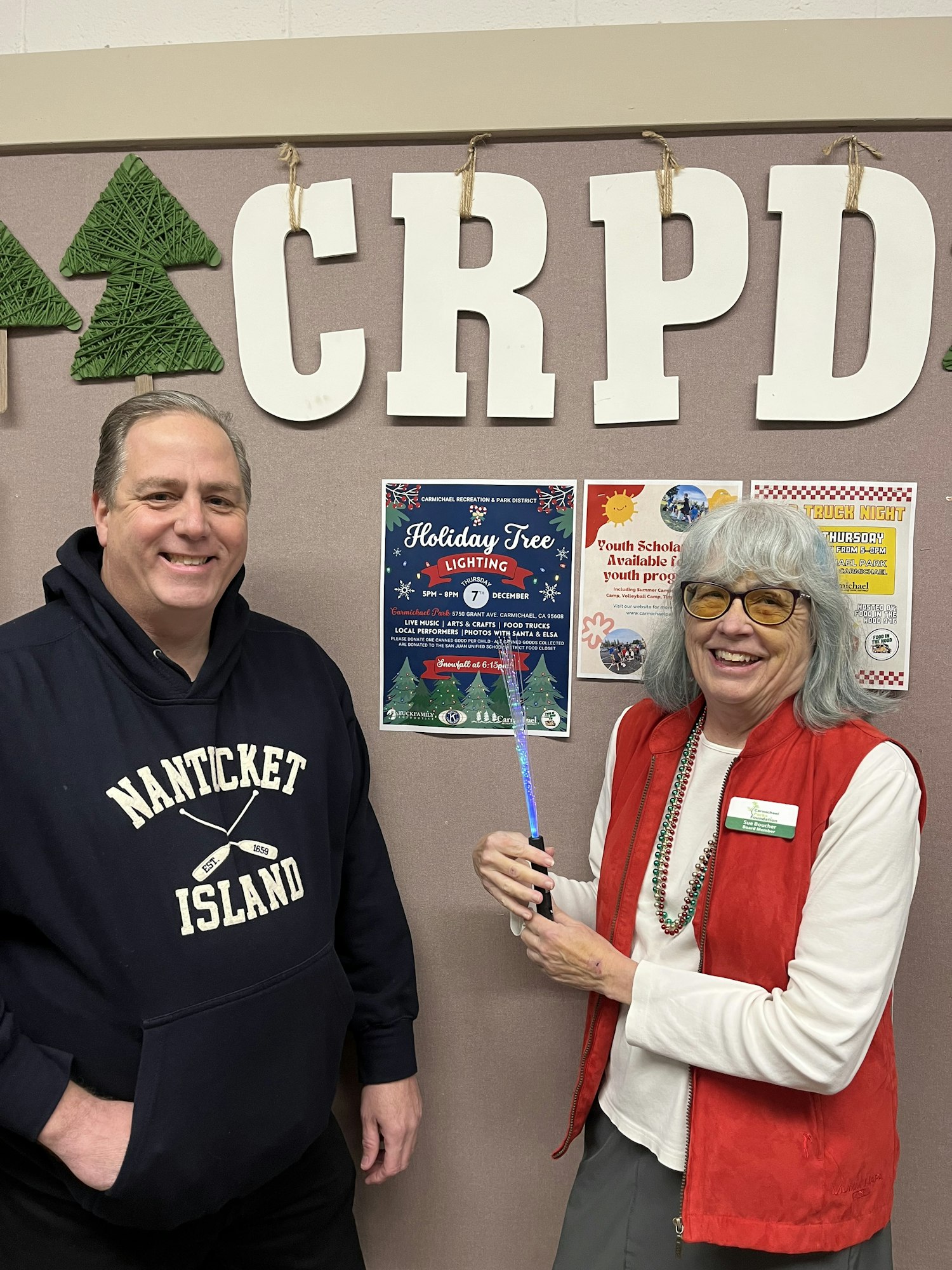 Two people posing, one standing with a pen, one sitting, near a bulletin board with the word "CRPD" and Christmas trees.