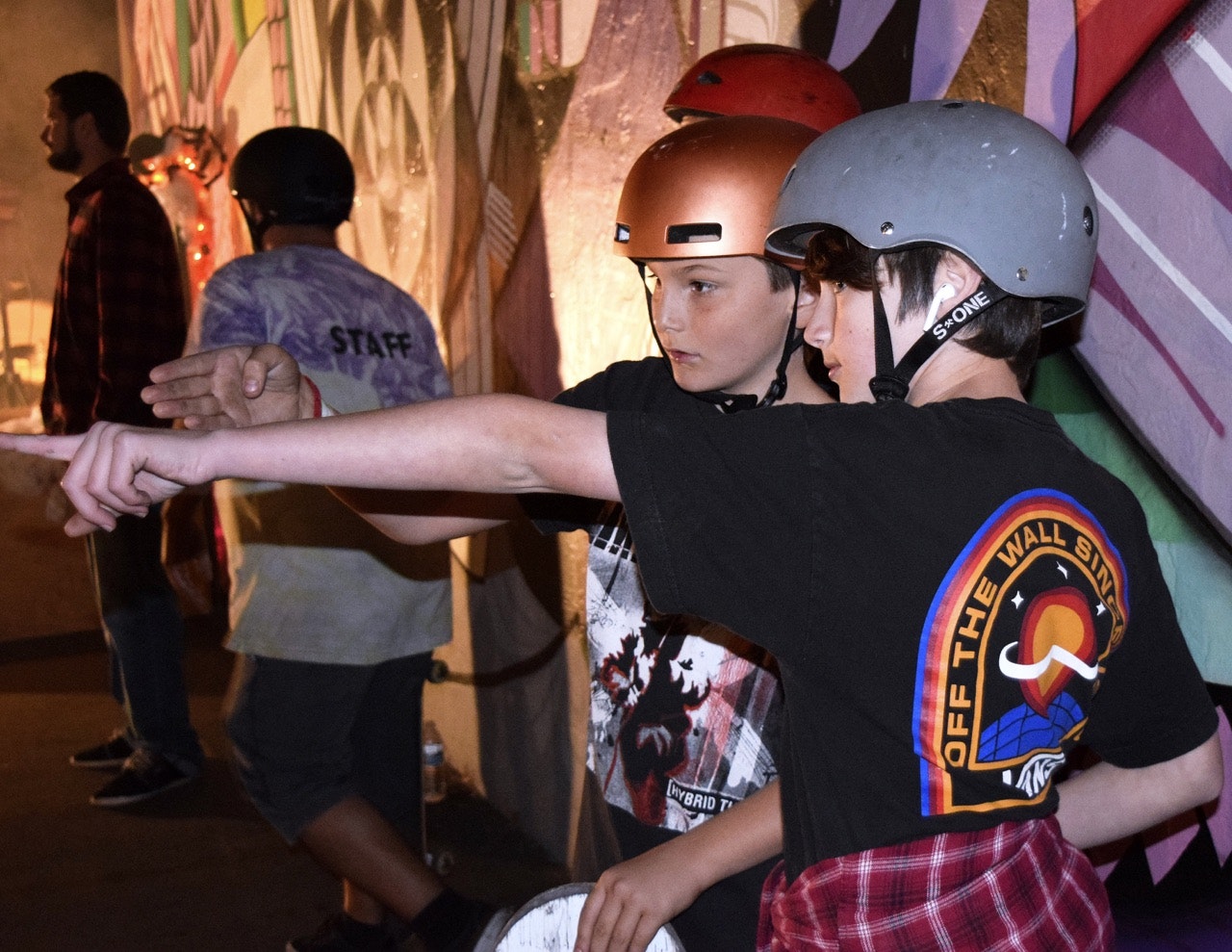 Two helmeted individuals are interacting, with one pointing and the other looking in the indicated direction; a person labeled 'STAFF' is in the background.