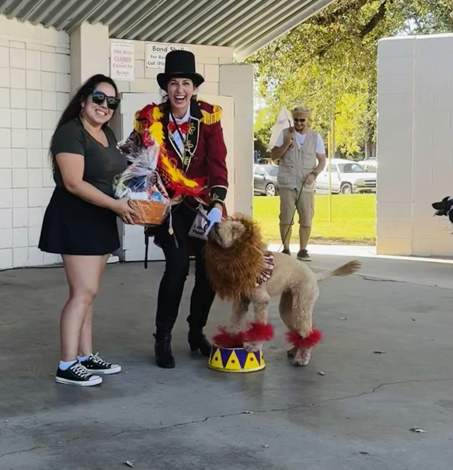Two people smiling; one holds a basket. A dog dressed as a lion stands on a platform. Another person and dog can be seen in the background.