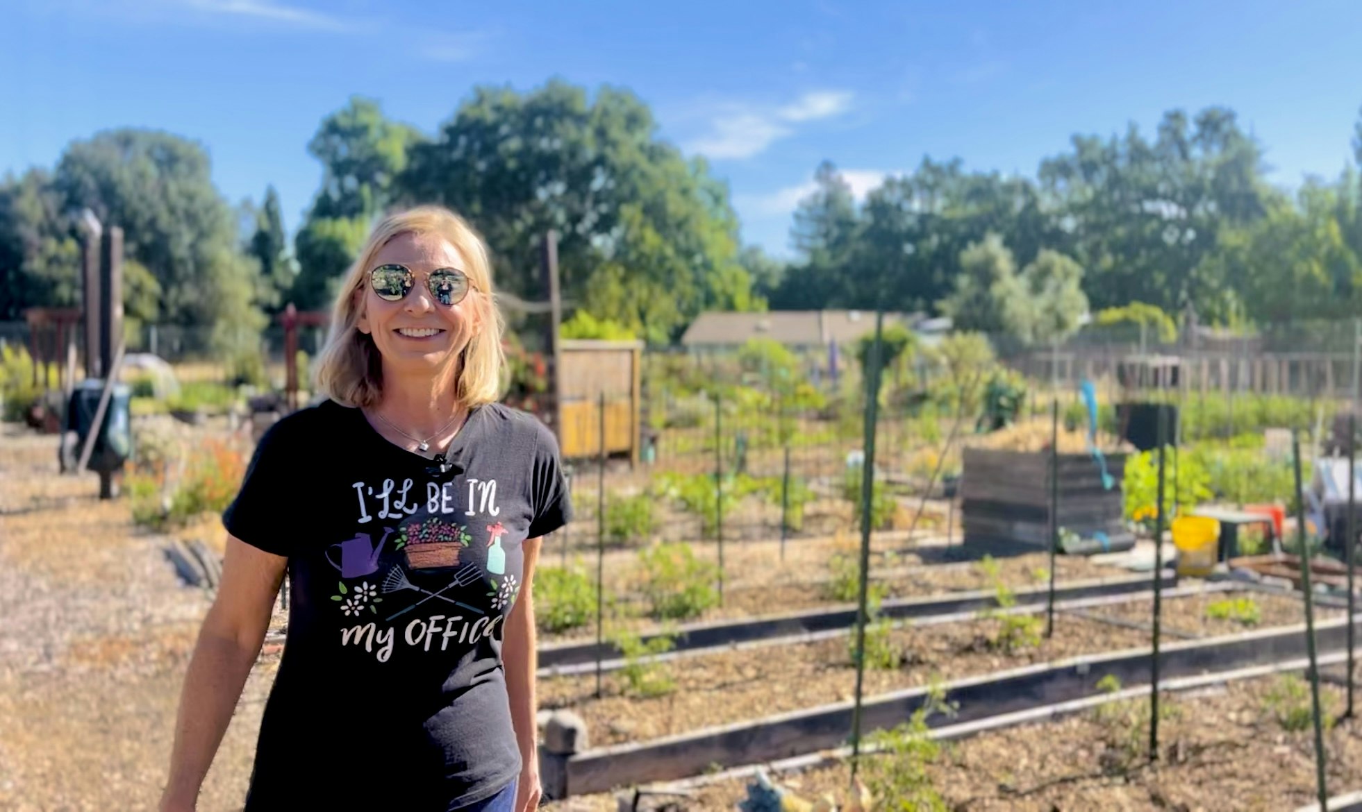 Woman in garden-themed t-shirt smiling in a sunny community garden.