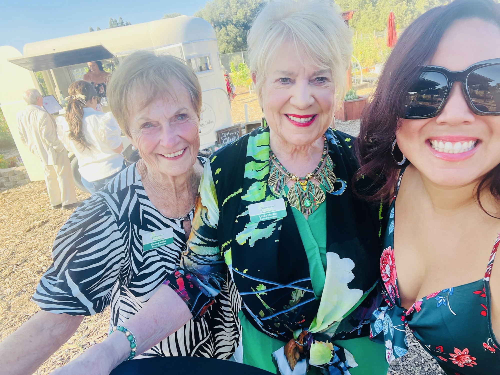 Three women smiling at an outdoor event with a food truck in the background.