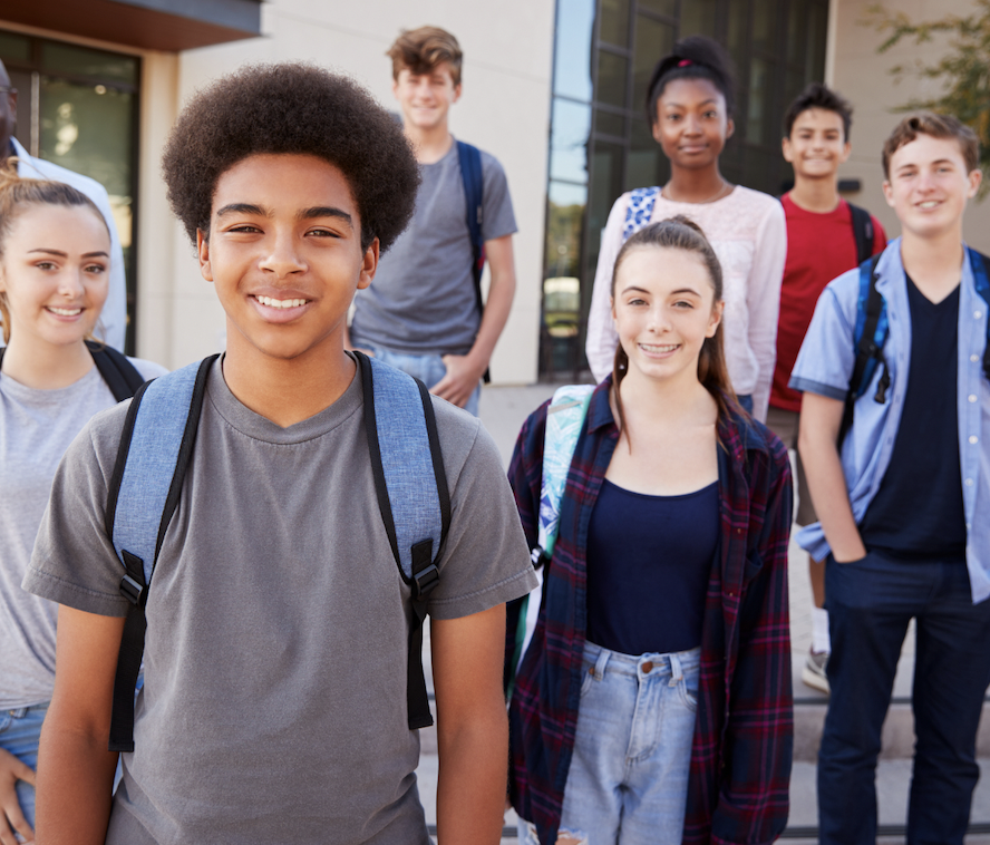 A group of diverse students with backpacks, smiling outside a school building.