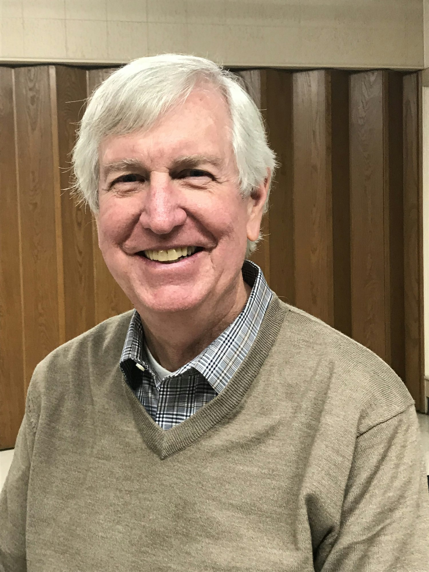 A smiling man with white hair wearing a v-neck sweater over a checkered shirt, standing in front of wood-paneled walls.