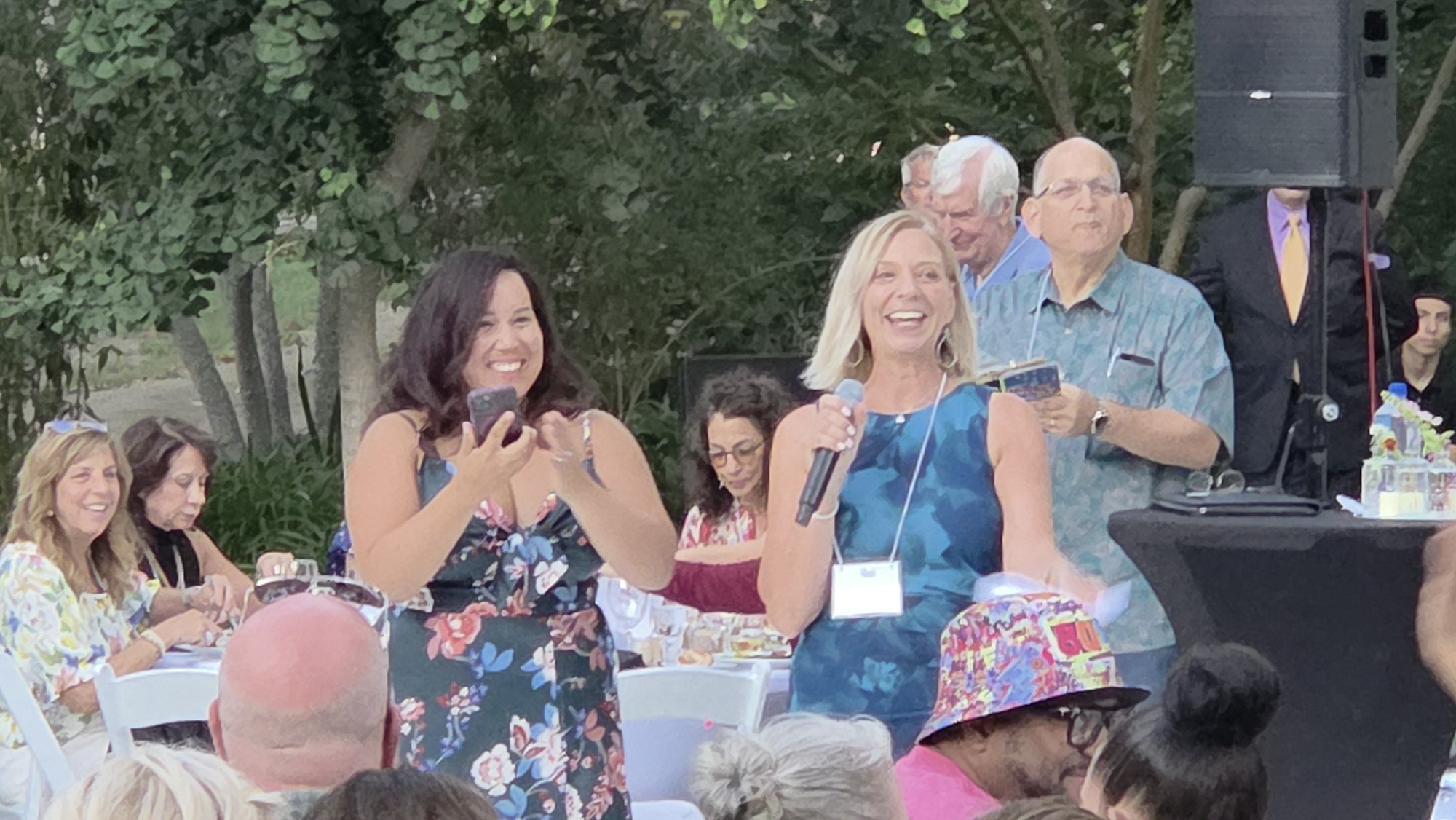 A woman with a microphone and another with a phone are smiling at an outdoor event. People are seated around them.