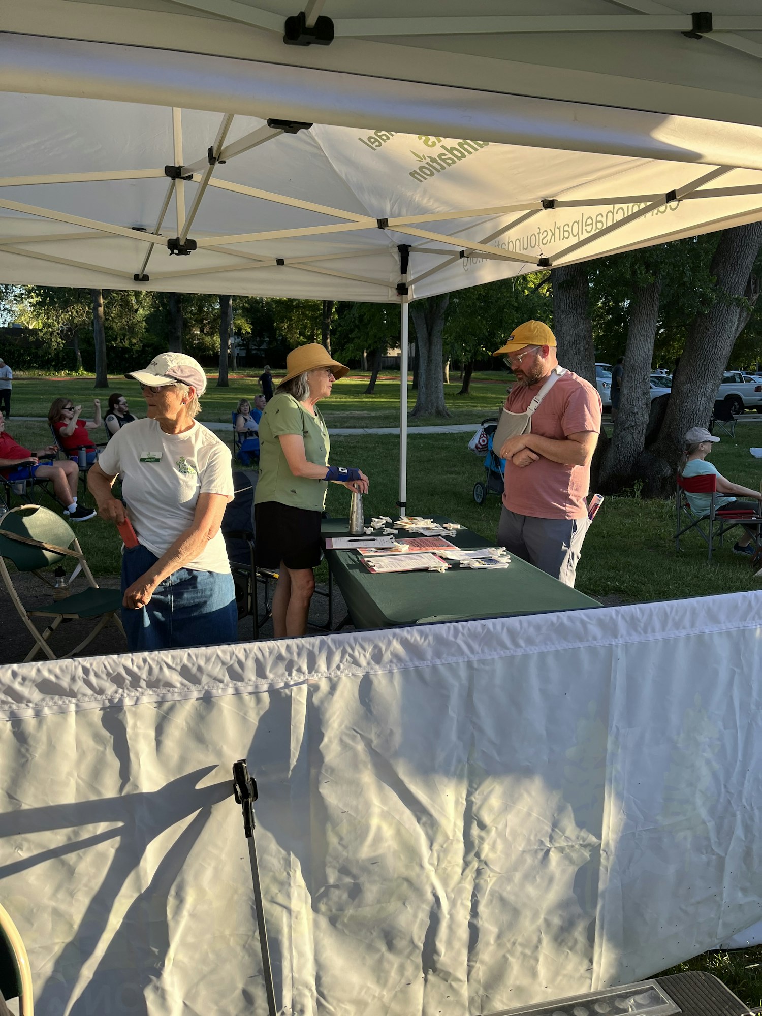 People under a tent with a table, in a park-like setting. Image is rotated; tops of trees are visible.