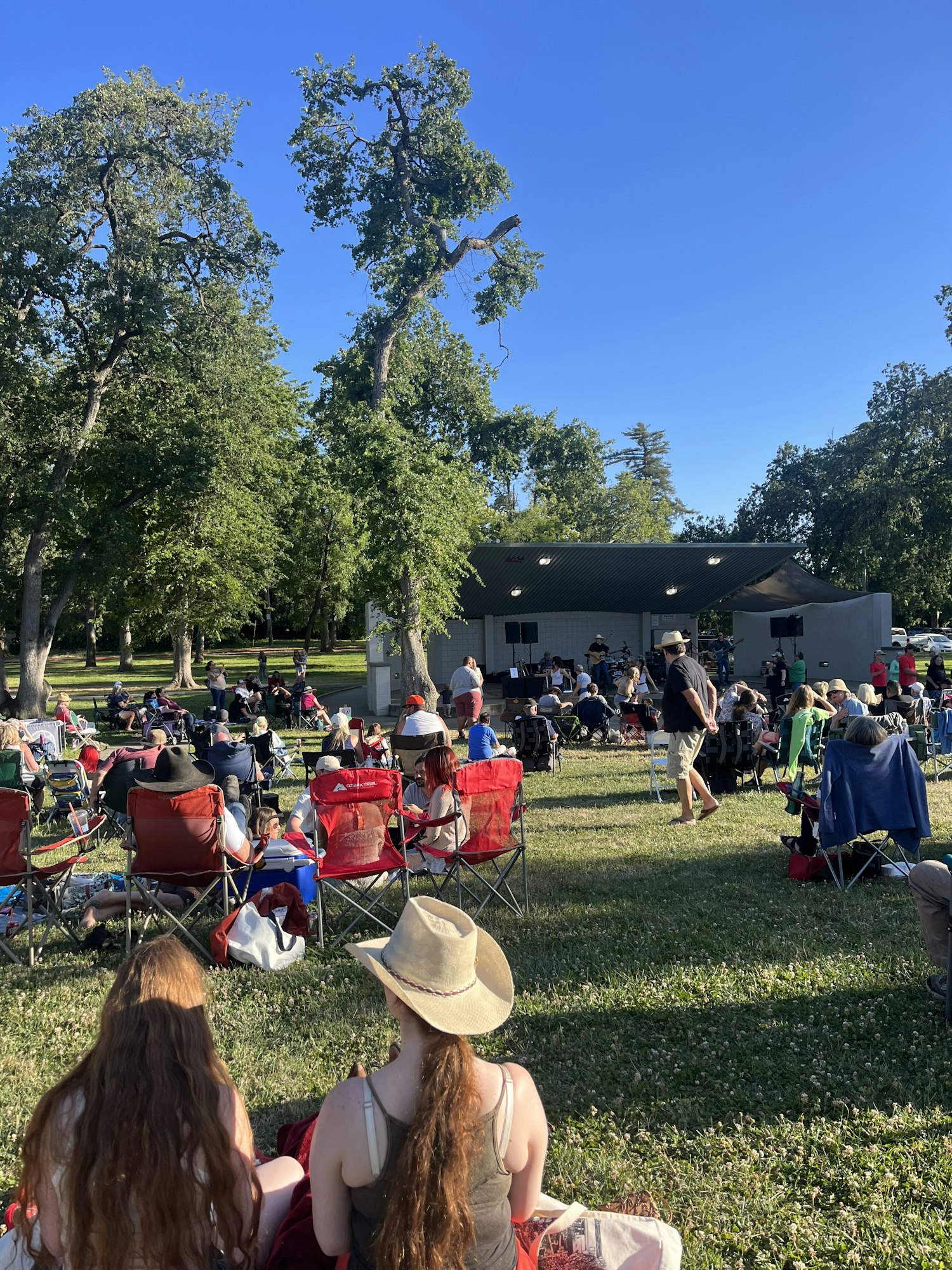Outdoor event with people sitting on lawn chairs, trees, and a building in the background.