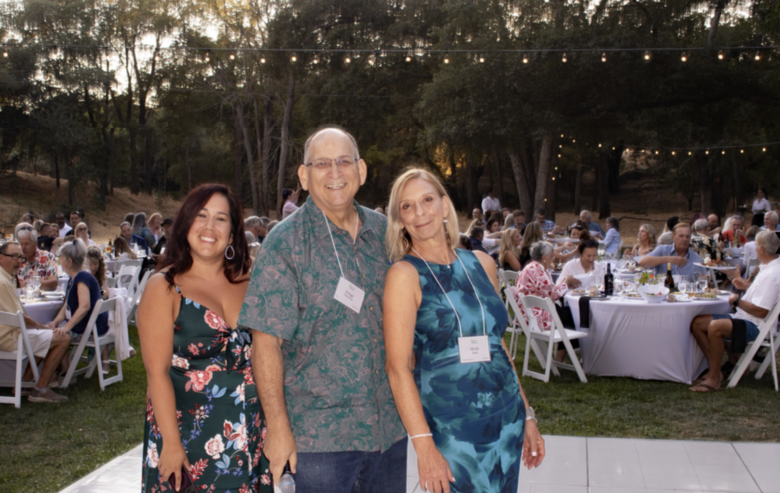 Three people pose at an outdoor event with string lights and guests seated at tables in the background.