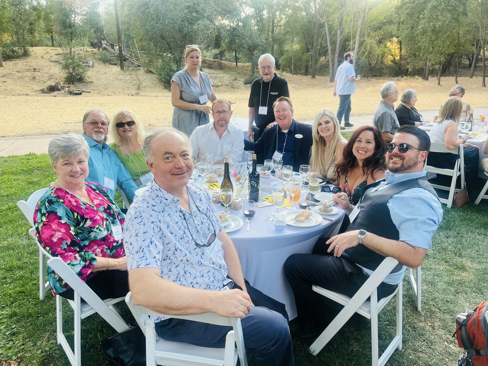 A group of people sitting around a table outdoors, enjoying a meal and smiling for a photo.