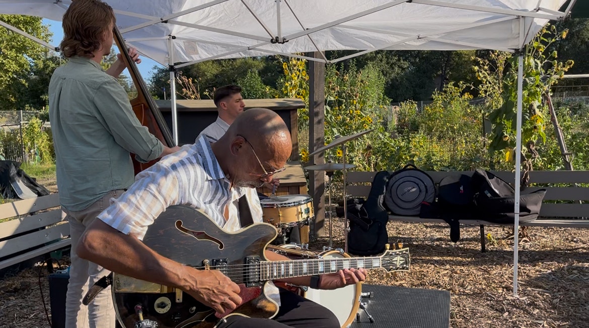 Three musicians perform outdoors: one plays guitar, another on double bass, and the third on drums, under a tent.