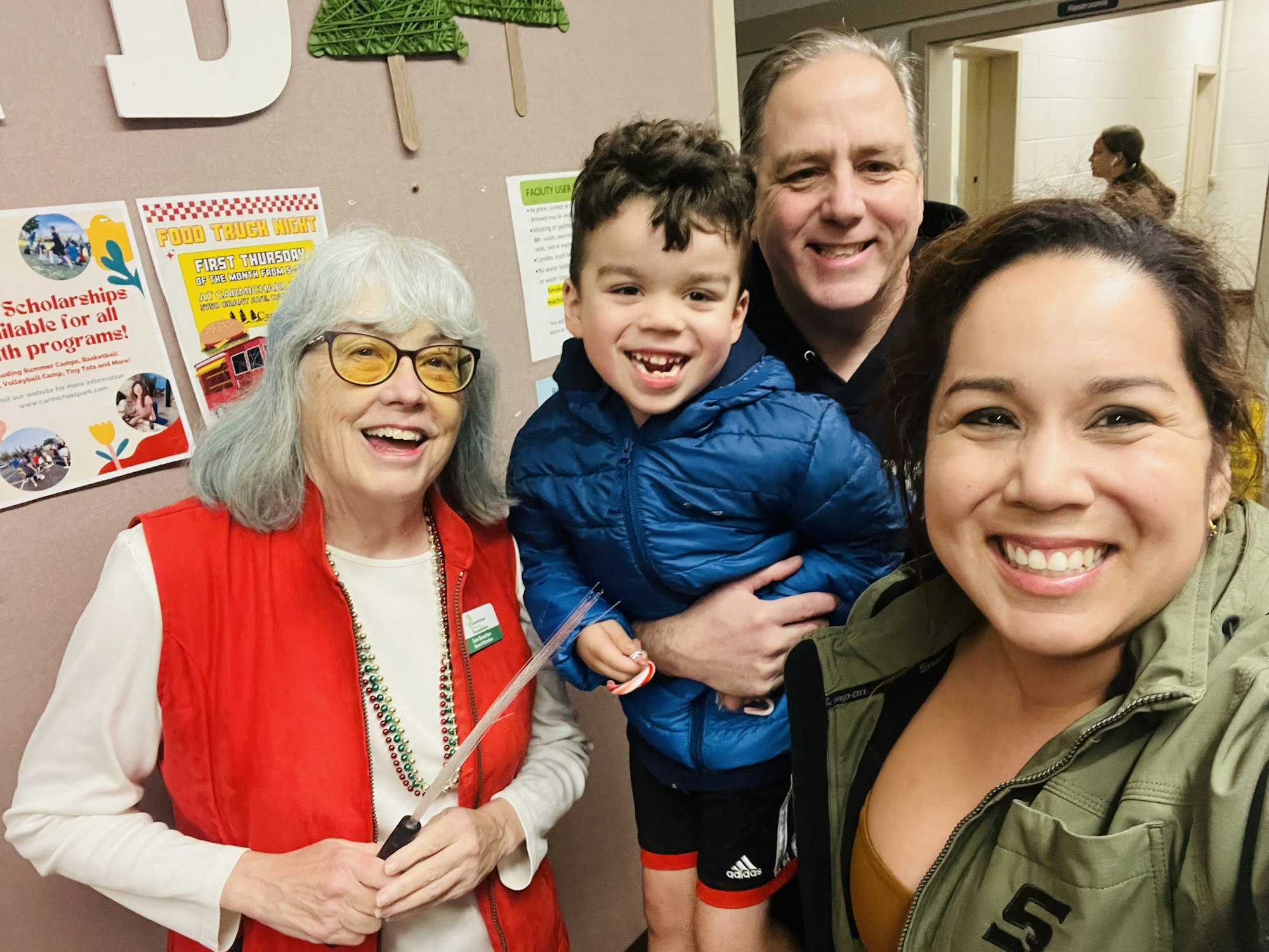 Four people smiling for a selfie; two adults, a child, and an older woman with a name tag. Background: bulletin board with flyers.