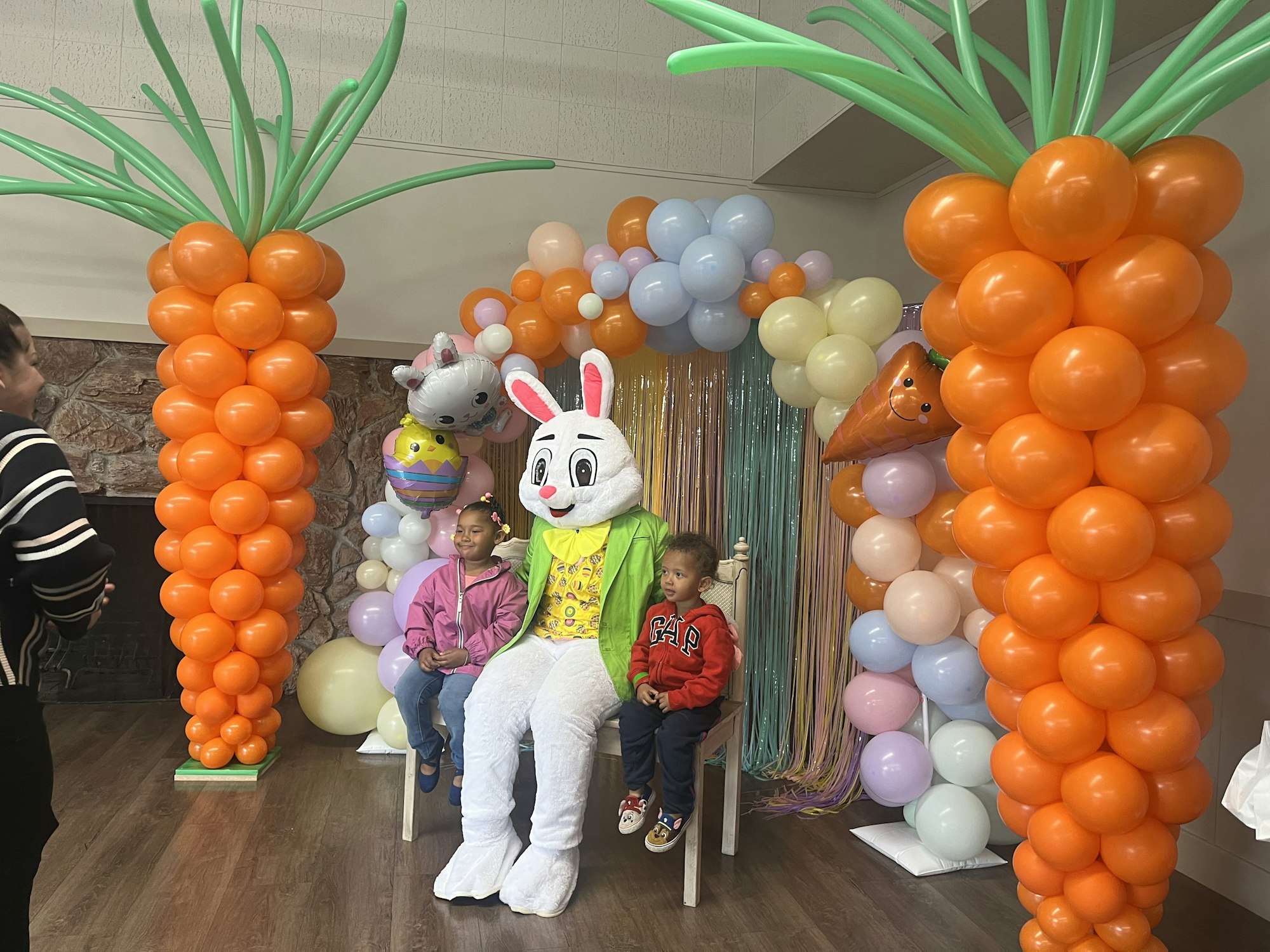 A person in a bunny costume is sitting with two kids among colorful balloon decorations.