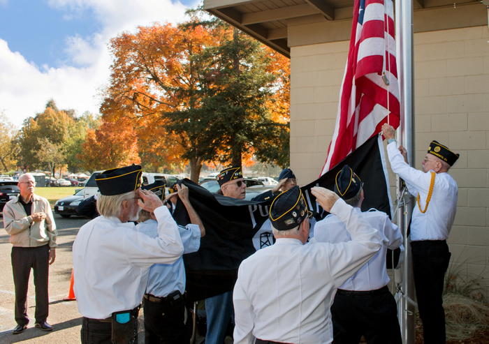Veterans in uniform raise the U.S. flag and a POW/MIA flag during a ceremony by a building with autumn trees in the background.