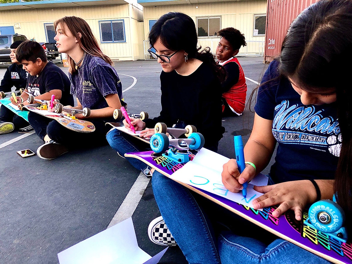 Group of kids sitting on ground with skateboards, writing on papers.