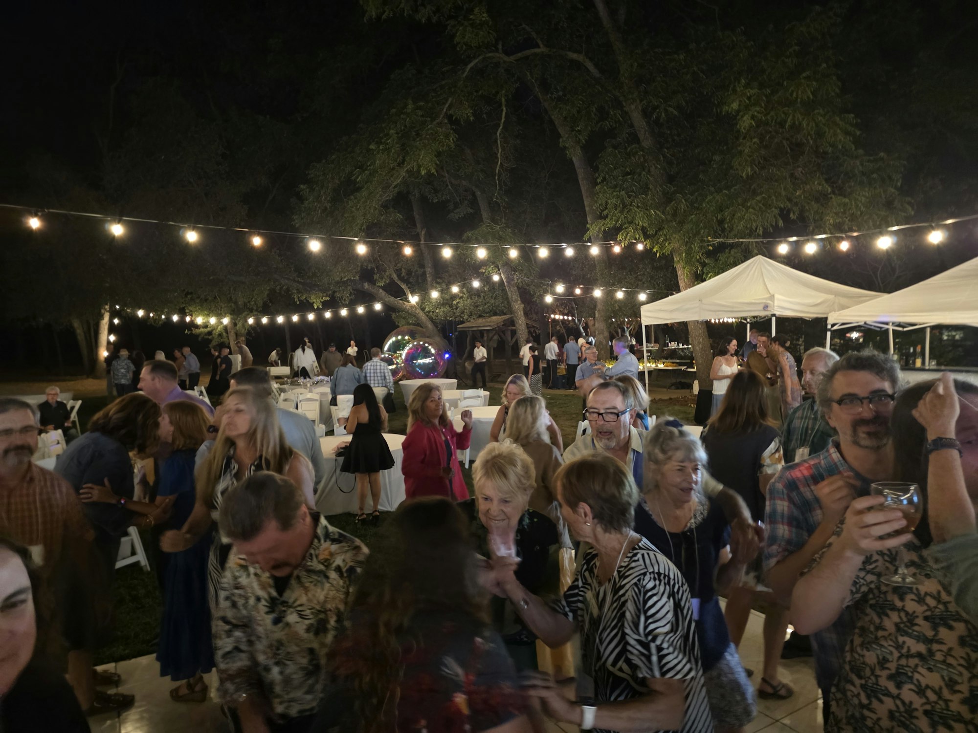 An outdoor gathering with people dancing under string lights, featuring a tent and trees in the background.
