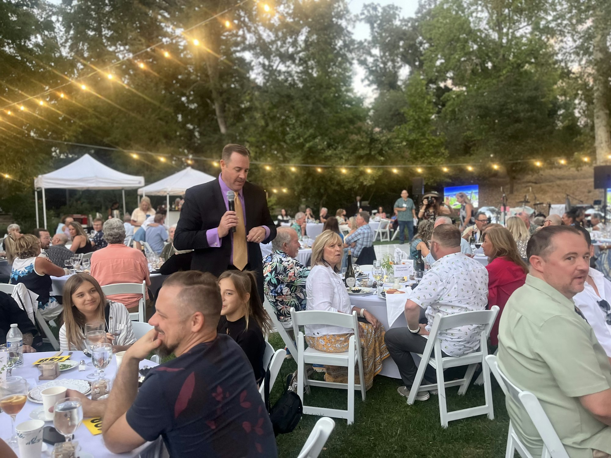 Outdoor gathering with people seated at tables under string lights, a speaker holding a microphone, and white tents in the background.
