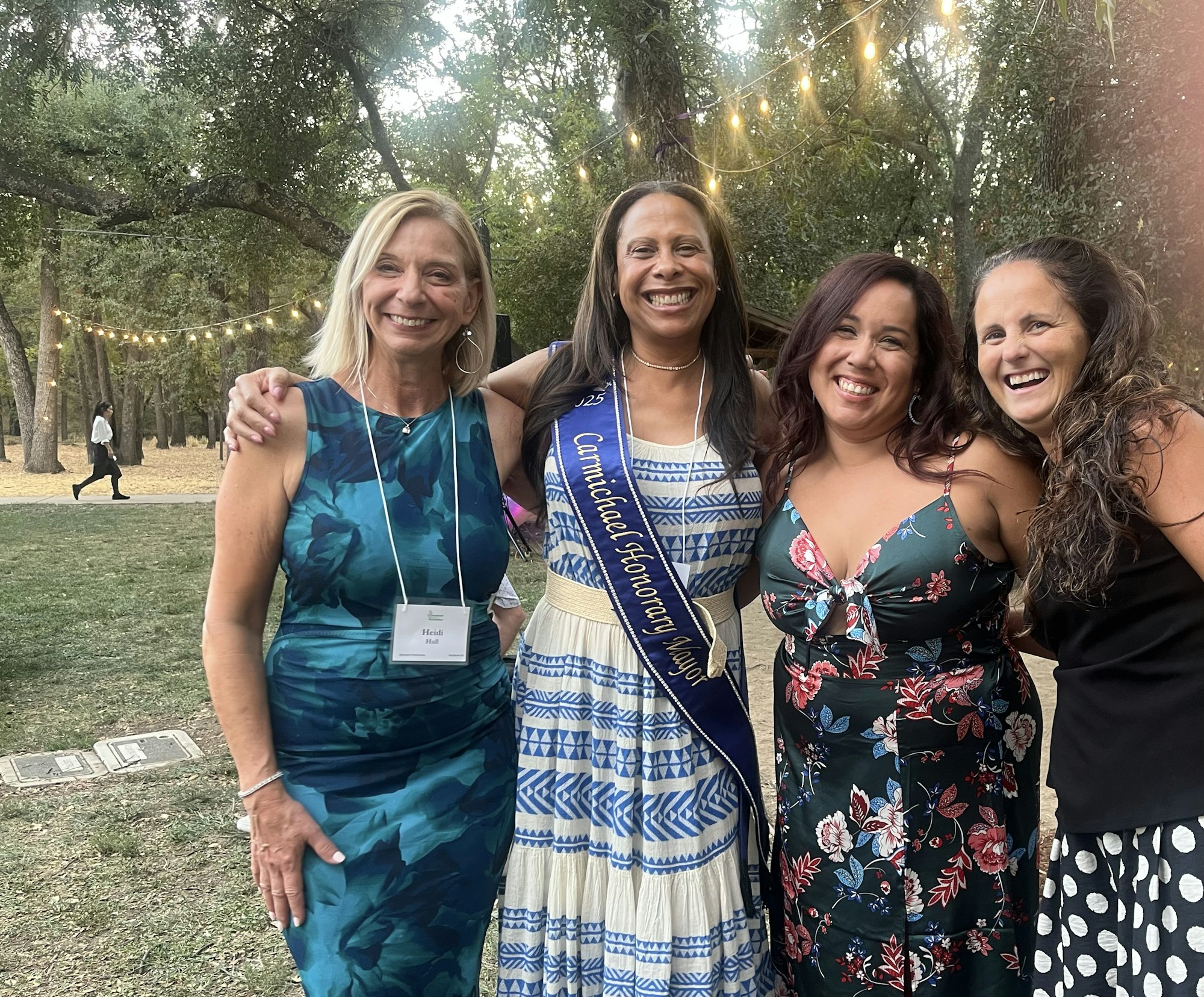 Four women smiling outdoors, one wearing a sash that says "Carnicadad Honorary Mayor." String lights and trees in the background.