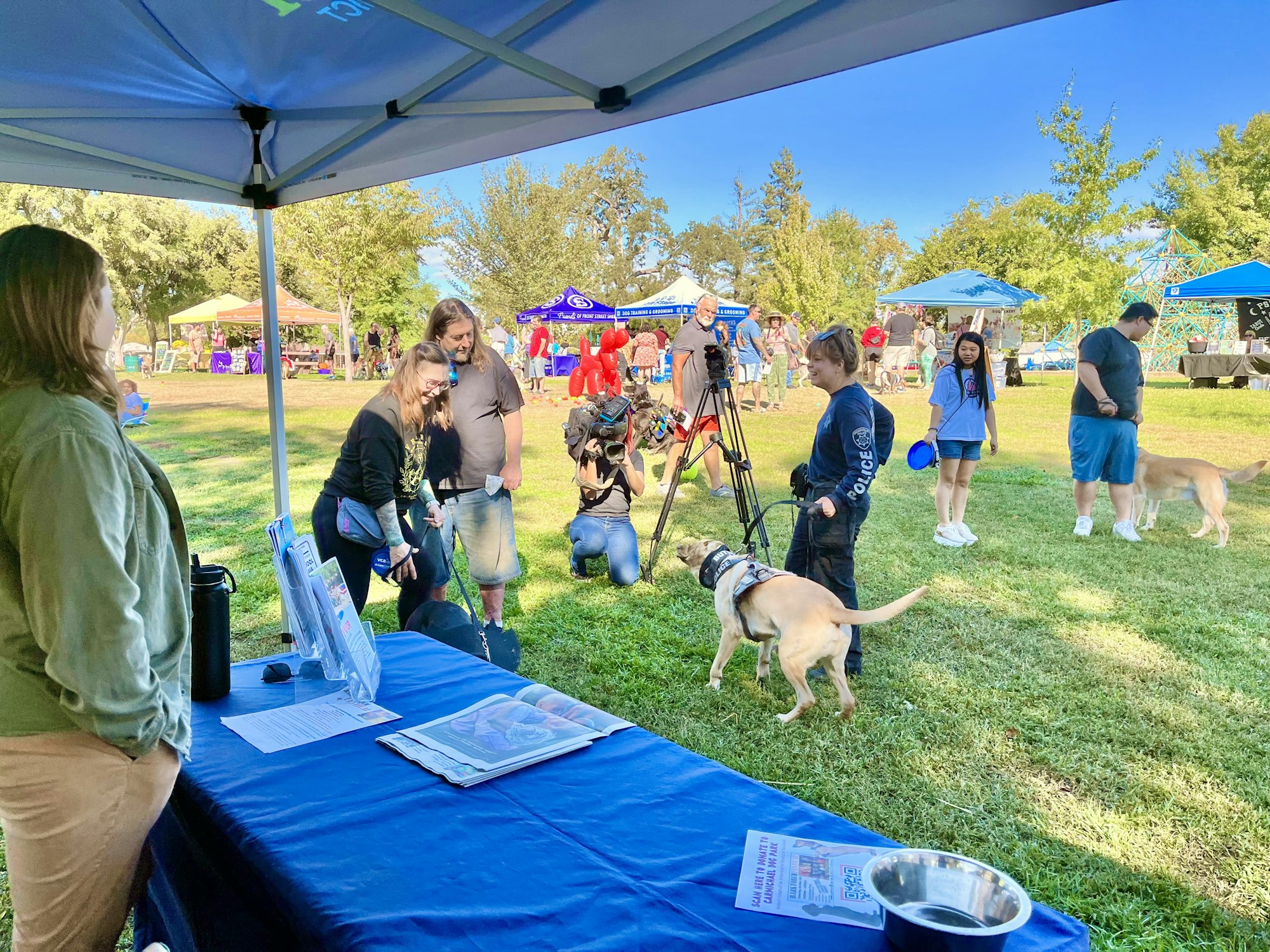 People and dogs at an outdoor event under a canopy, with tents and informational displays in a park setting.