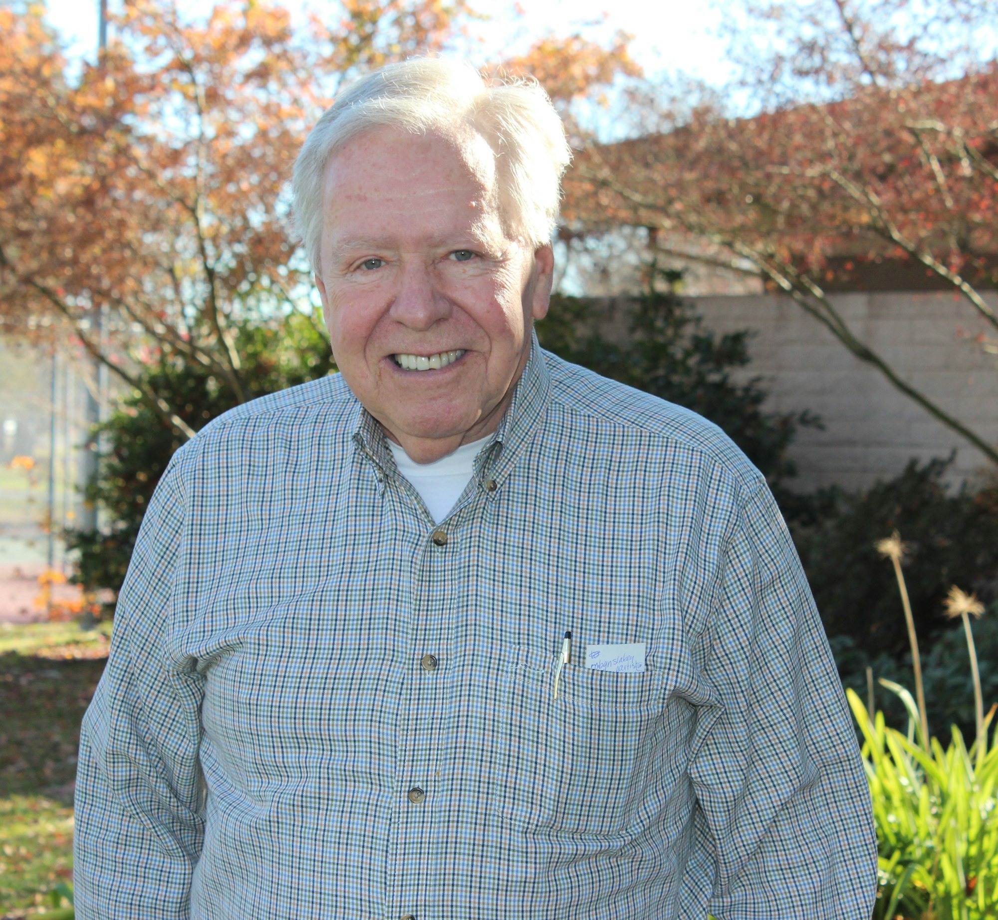 Smiling man outdoors, wearing a checked shirt, with autumn trees in the background.