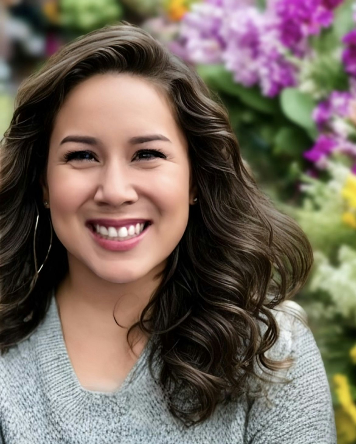 A smiling woman with curly hair, wearing earrings and a grey sweater, with blurred purple flowers in the background.